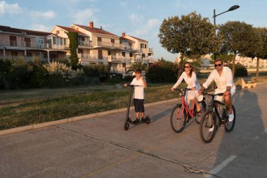 Mutlu bir aile deniz kenarında güzel bir sabah geçiriyorlar, ebeveynler bisiklete biniyor ve oğulları elektrikli scooter kullanıyor. Seçici odak 
