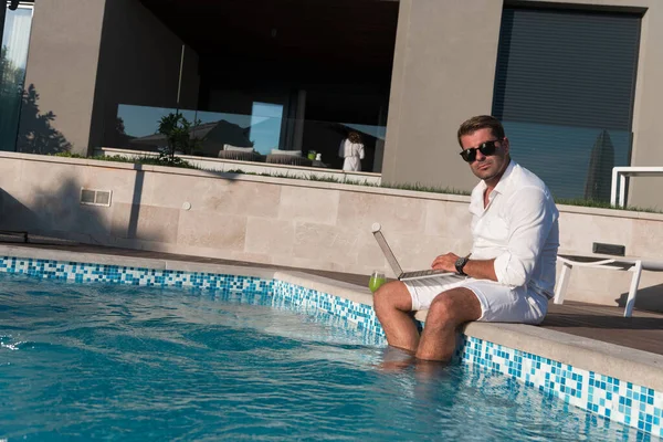 Um homem idoso moderno desfruta da piscina enquanto trabalha em seu laptop ao lado de uma casa de luxo moderna. Foco seletivo — Fotografia de Stock