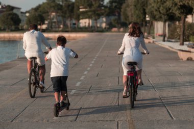Mutlu bir aile deniz kenarında güzel bir sabah geçiriyorlar, ebeveynler bisiklete biniyor ve oğulları elektrikli scooter kullanıyor. Seçici odak 