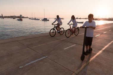 Mutlu bir aile deniz kenarında güzel bir sabah geçiriyorlar, ebeveynler bisiklete biniyor ve oğulları elektrikli scooter kullanıyor. Seçici odak 