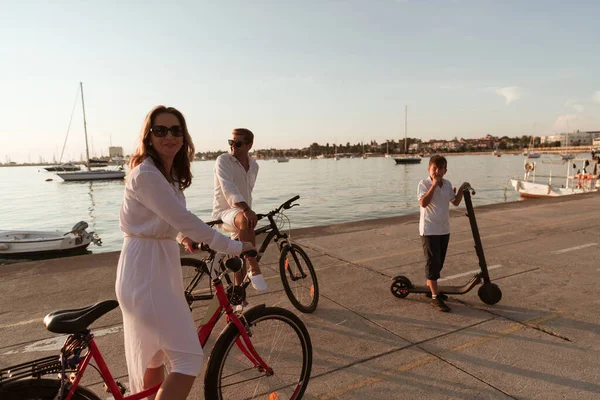 Szczęśliwa rodzina ciesząca się pięknym porankiem nad morzem, rodzice jeżdżący na rowerze i ich syn jeżdżący na skuterze elektrycznym. Skupienie selektywne — Zdjęcie stockowe