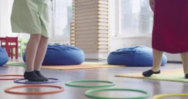 Küçük anaokulu öğrencileri ve bayan öğretmenleri sınıfın içinde egzersiz yapıyorlar. Hula hoop çemberinin üzerinden zıplarken yerde.