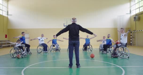 El selector del equipo de baloncesto con discapacidad se coloca delante de los jugadores y les muestra los ejercicios de estiramiento antes del inicio del entrenamiento — Vídeo de stock