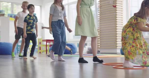 教室内の床の上に女性教師を持つ小さな保育園の子供たちは、運動をします。床の上にフラフープサークルトラック上でジャンプ. — ストック動画