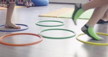 Küçük anaokulu öğrencileri ve bayan öğretmenleri sınıfın içinde egzersiz yapıyorlar. Hula hoop çemberinin üzerinden zıplarken yerde.