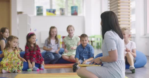Storytime elementary or primary school teacher reading a story to a group of children in a daycare center. — Vídeo de Stock
