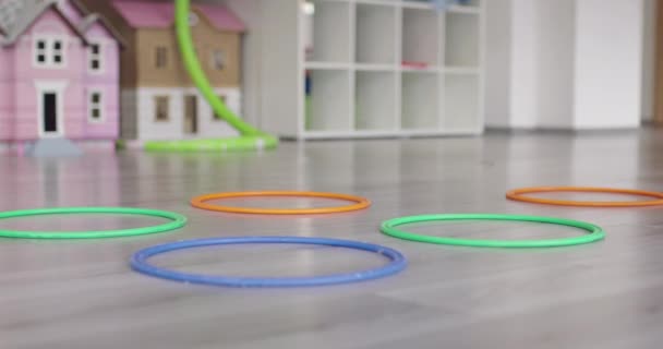 Små förskolebarn med kvinnlig lärare på golvet inomhus i klassrummet, motionerar. Hoppa över hula båge cirklar spår på golvet. — Stockvideo