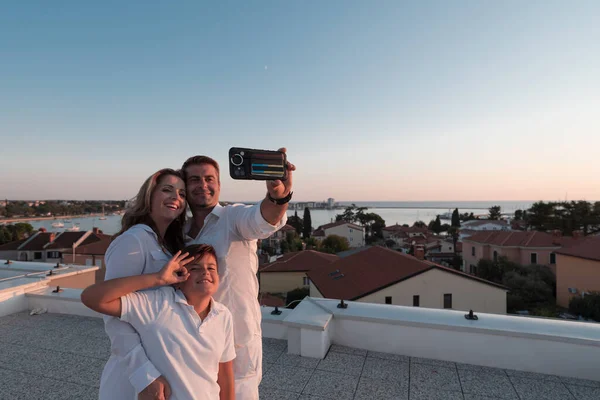 Gelukkig gezin neemt een selfie met een smartphone op het dak van hun huis bij zonsondergang. Selectieve focus — Stockfoto