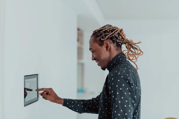 Sonriente hombre afroamericano utilizando un moderno sistema de hogar inteligente, controlador en la pared, joven positivo cambiando la temperatura en el termostato o activando la alarma de seguridad en el apartamento —  Fotos de Stock