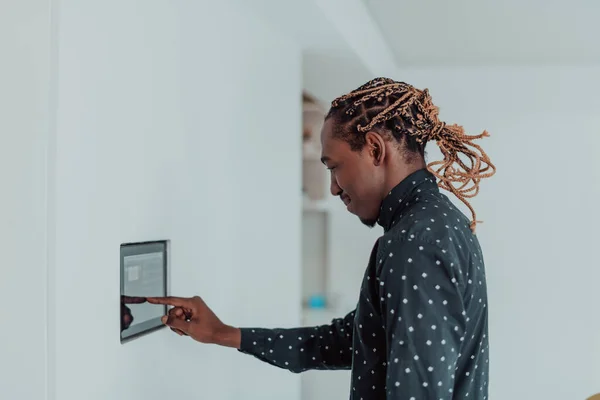 Lächelnder Afroamerikaner mit modernem Smart-Home-System, Controller an der Wand, positiver junger Mann schaltet Thermostat ein oder aktiviert Sicherheitsalarm in Wohnung — Stockfoto