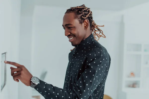 Sonriente hombre afroamericano utilizando un moderno sistema de hogar inteligente, controlador en la pared, joven positivo cambiando la temperatura en el termostato o activando la alarma de seguridad en el apartamento —  Fotos de Stock