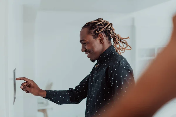 Sonriente hombre afroamericano utilizando un moderno sistema de hogar inteligente, controlador en la pared, joven positivo cambiando la temperatura en el termostato o activando la alarma de seguridad en el apartamento —  Fotos de Stock
