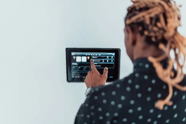 Lächelnder Afroamerikaner mit modernem Smart-Home-System, Controller an der Wand, positiver junger Mann schaltet Thermostat ein oder aktiviert Sicherheitsalarm in Wohnung — Stockfoto