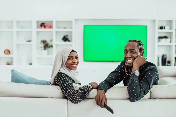 Pareja africana sentada en un sofá viendo televisión juntos Pantalla verde croma Mujer vestida con ropa islámica de Hijab —  Fotos de Stock