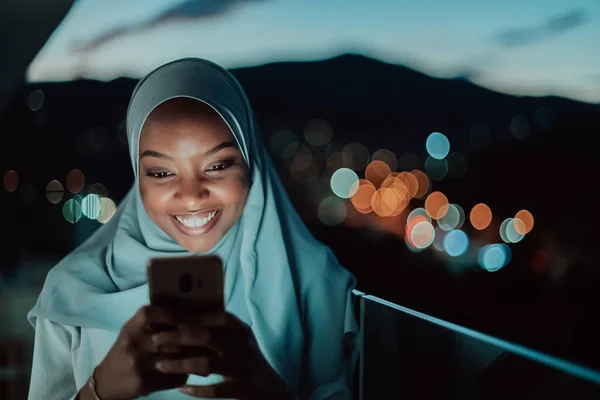 Młoda muzułmanka nosząca chustę na miejskiej ulicy w nocy smsująca na smartfonie ze światłem miasta bokeh w tle. — Zdjęcie stockowe