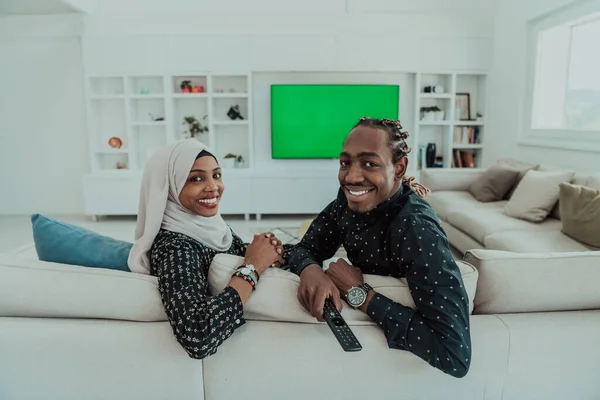 ソファに座っているアフリカのカップルテレビを一緒に見るクロマ緑の画面イスラム教のヒジャブを身に着けている女性服 — ストック写真