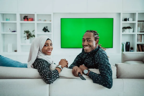 Pareja africana sentada en un sofá viendo televisión juntos Pantalla verde croma Mujer vestida con ropa islámica de Hijab —  Fotos de Stock