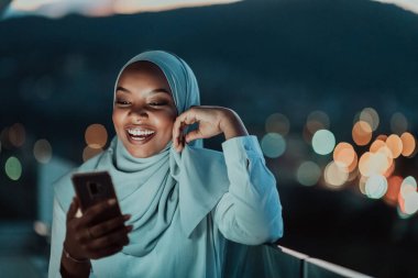 Gece şehir caddesinde fular takan genç Müslüman bir kadın arka planda Bokeh şehir ışığı olan bir telefondan mesaj atıyor.. 