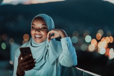 Young Muslim woman wearing scarf veil on urban city street at night texting on a smartphone with bokeh city light in the background.  clipart