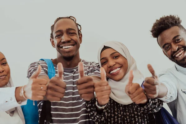 Retrato grupal de estudiantes africanos felices parados juntos sobre un fondo blanco y mostrando un cartel aceptable con pulgares hacia arriba a las niñas que usan la moda tradicional sudanesa musulmana de hiyab —  Fotos de Stock
