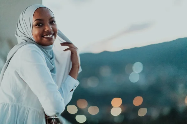 Afrikanische Muslimin in der Nacht auf einem Balkon, der in die Kamera lächelt, mit Bokeh-Lichtern im Hintergrund. — Stockfoto