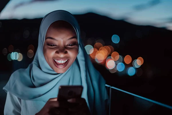 Giovane donna musulmana indossa velo sciarpa sulla strada urbana della città di notte sms su uno smartphone con bokeh luce della città sullo sfondo. — Foto Stock