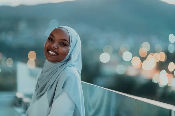 Afrikanische Muslimin in der Nacht auf einem Balkon, der in die Kamera lächelt, mit Bokeh-Lichtern im Hintergrund. — Stockfoto