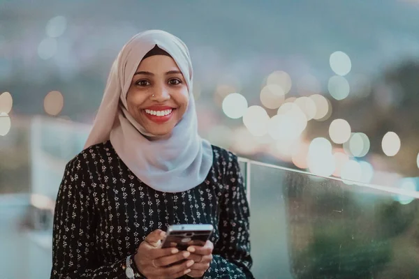 Mujer musulmana joven con velo de bufanda en la calle urbana de la ciudad por la noche mensajes de texto en un teléfono inteligente con luz de la ciudad bokeh en el fondo. — Foto de Stock
