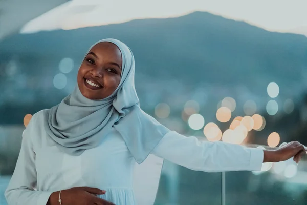 Afrikanische Muslimin in der Nacht auf einem Balkon, der in die Kamera lächelt, mit Bokeh-Lichtern im Hintergrund. — Stockfoto