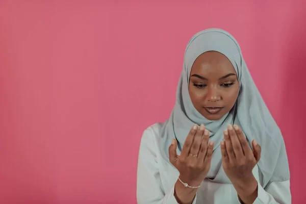 Moderne afrikanische Muslimin betet traditionell zu Gott, hält die Hände in Gebetsgesten, trägt traditionelle weiße Kleidung, hat einen ernsten Gesichtsausdruck, isoliert über einem rosafarbenen Plastikhintergrund — Stockfoto