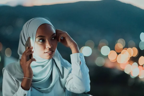 Jeune femme musulmane portant un voile d'écharpe dans la rue de la ville urbaine la nuit textos sur un smartphone avec la lumière de la ville bokeh en arrière-plan. — Photo