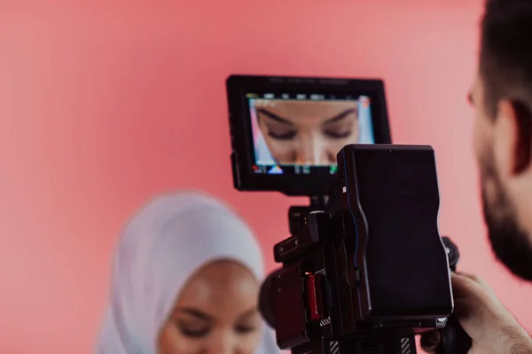 Videographer in digital studio recording video on professional camera by shooting female Muslim woman wearing hijab scarf plastic pink background. — Stock Photo, Image