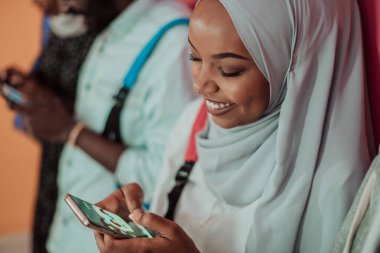 Bir grup Afrikalı Müslüman öğrenci sarı bir arkaplan önünde dururken akıllı telefon kullanıyor.