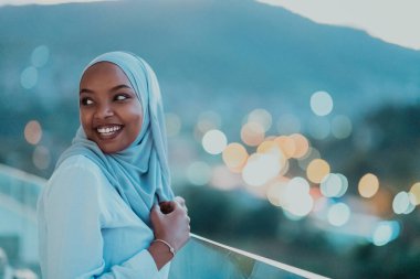 Geceleyin Afrikalı Müslüman bir kadın balkonda kameraya gülümsüyor. Arka planda bokeh ışıkları var..