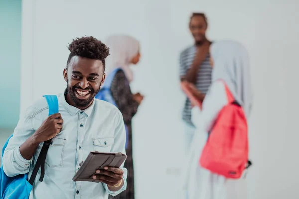 Style de vie universitaire beau jeune homme étudiant africain tenant une tablette informatique et souriant tout en se tenant contre l'université avec ses amis ont une réunion d'équipe en arrière-plan. Haute qualité — Photo