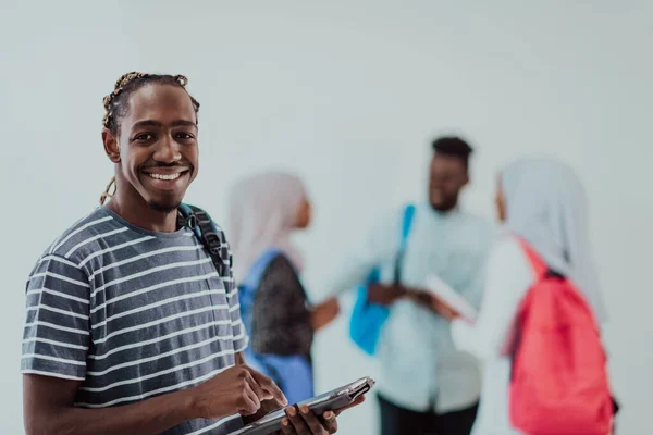Estilo de vida universitario joven estudiante africano guapo sosteniendo una tableta y sonriendo mientras se enfrenta a la universidad con sus amigos tienen una reunión de equipo en el fondo. Alta calidad —  Fotos de Stock