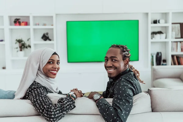 Αφρικανικό ζευγάρι κάθεται στον καναπέ βλέποντας τηλεόραση μαζί Chroma πράσινη οθόνη γυναίκα φορώντας ισλαμικά ρούχα μαντίλα — Φωτογραφία Αρχείου