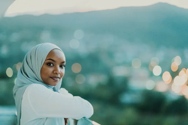 非洲穆斯林妇女在夜间的阳台上微笑着看着摄像机，背后是城市的灯火通明. — 图库照片