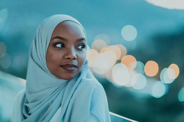 Afrikai muszlim nő az éjszaka egy erkélyen mosolyog a kamera a város bokeh fények a háttérben. — Stock Fotó