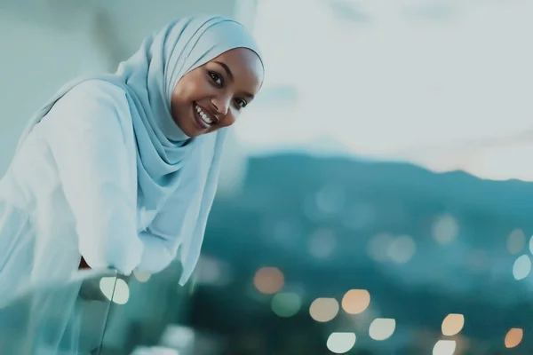 Africký muslim žena v noci na balkóně s úsměvem na kameru s městem bokeh světla v pozadí. — Stock fotografie