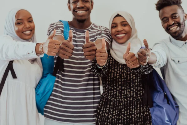 Retrato grupal de estudiantes africanos felices parados juntos sobre un fondo blanco y mostrando un cartel aceptable con pulgares hacia arriba a las niñas que usan la moda tradicional sudanesa musulmana de hiyab —  Fotos de Stock