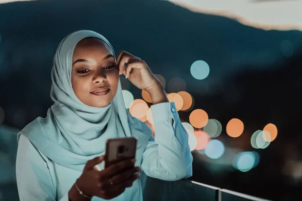 Młoda muzułmanka nosząca chustę na miejskiej ulicy w nocy smsująca na smartfonie ze światłem miasta bokeh w tle. — Zdjęcie stockowe