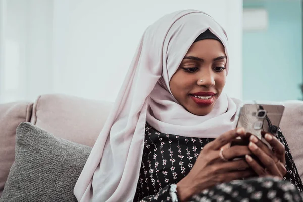 Wanita Afrika menggunakan smartphone ketika duduk di sofa di rumah mengenakan pakaian jilbab. — Stok Foto
