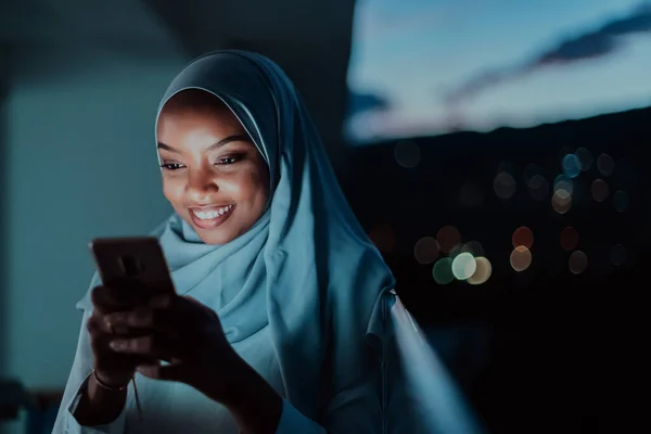 Wanita muslim muda mengenakan kerudung syal di jalan kota pada malam hari SMS di smartphone dengan lampu bokeh kota di latar belakang. — Stok Foto