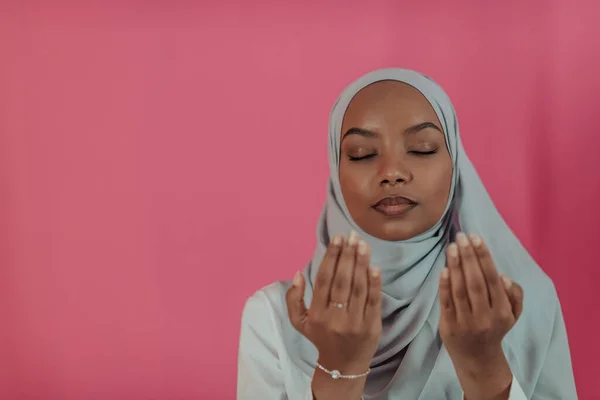 Mulher muçulmana africana moderna faz oração tradicional a Deus, mantém as mãos em gesto de oração, veste roupas brancas tradicionais, tem expressão facial séria, isolado sobre fundo de plástico rosa — Fotografia de Stock