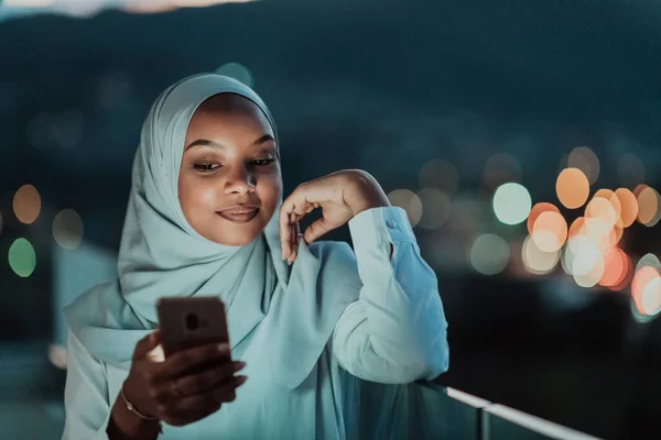 Mujer musulmana joven con velo de bufanda en la calle urbana de la ciudad por la noche mensajes de texto en un teléfono inteligente con luz de la ciudad bokeh en el fondo. —  Fotos de Stock