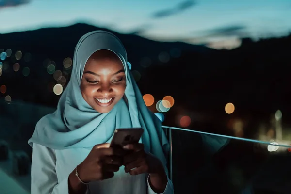 O tânără musulmană poartă văl de eșarfă pe strada urbană a orașului pe timp de noapte, trimițând mesaje text pe un smartphone cu lumina orașului bokeh în fundal . — Fotografie, imagine de stoc