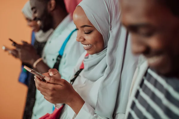 A group of African Muslim students use smartphones while standing in front of a yellow background — Stock Photo, Image