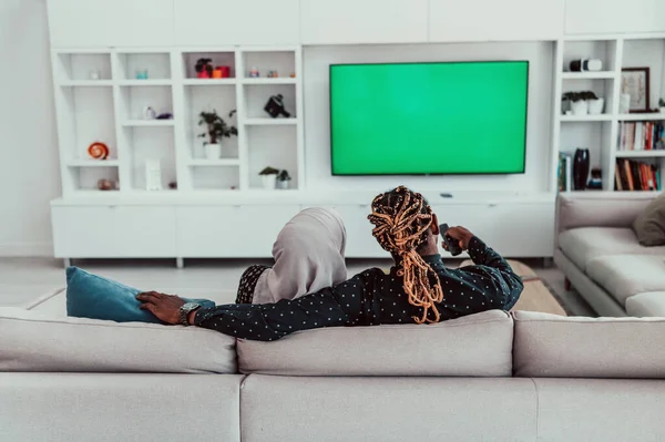 Африканская пара, сидящая на диване и смотрящая вместе телевизор, хрома зеленый экран женщина в исламском хиджабе Одежда — стоковое фото