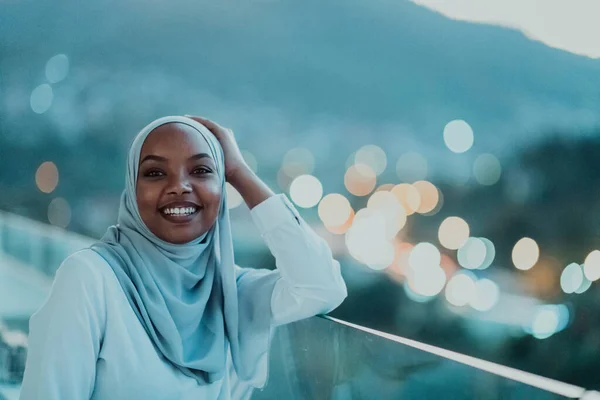Wanita Muslim Afrika di malam hari di balkon tersenyum di kamera dengan lampu bokeh kota di latar belakang. — Stok Foto